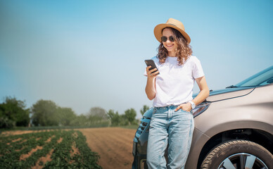 Happy cheerful young woman in hat and sunglasses with smartphone standing by the car
