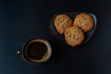 Top-down dark still life with a cup of espresso and oatmeal cookies on a black saucer