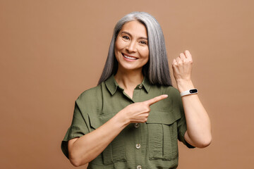 Happy positive woman with grey hair in pointing finger on watch on her wrist looking at camera with toothy smile. Look at time. Indoor studio shot isolated on brown background