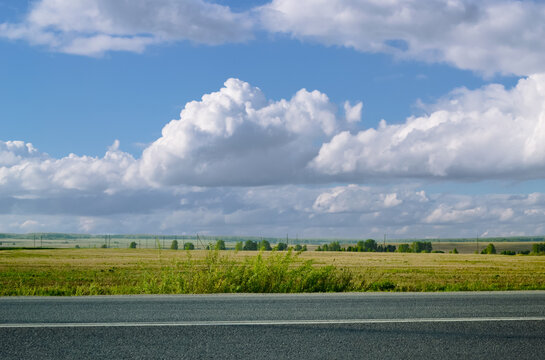 Majestic clouds over an asphalt road, bushes and a field with trees