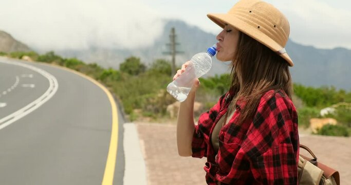 woman drinking water while resting on the rock during the travel in the mountains. A young caucasian woman in the mountains with a backpack stands and drinks water from a personal refillable bottle