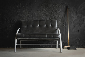 Black dirty sofa, broom and dustpan on the black wall background.