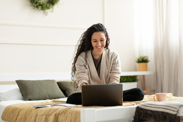 Joyful young lady sitting cross legged on bed, using laptop for online work or education at home,...