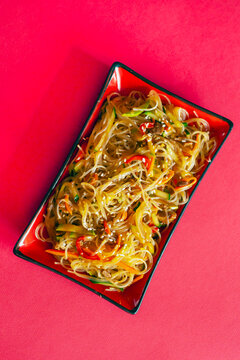 Traditional Korean starch noodles chapche with vegetables and beef in a red rectangular plate on a red background, vertical shot.