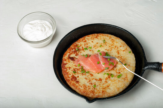traditional Swiss dish is a potato pancake in a frying pan with pieces of red salmon fish, decorated with green onions. there is a gravy boat with white cream sauce nearby.