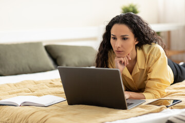 Naklejka premium Serious young woman using laptop, working or studying online, having business meeting or webinar, lying on bed at home