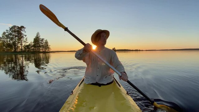 Young man in a straw hat and linen shirt paddles with a yellow oar on a yellow kayak on a calm forest lake at sunset on a summer evening in slow motion.