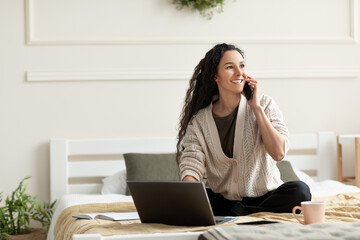 Cheerful young woman with laptop pc communicating on cellphone, sitting on bed at home, free space