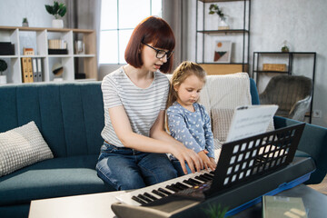 Mom and little happy girl in music therapy by playing piano on music room. Teacher helping young female pupil in piano lesson. Relaxing at home.