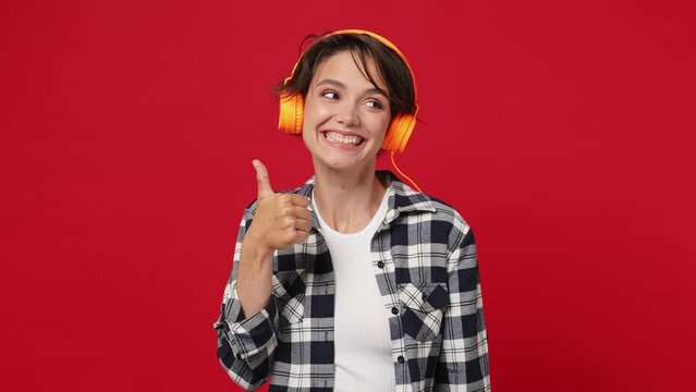 Vivid cheerful woman 20s she wearing basic white t-shirt checkered shirt listen music in headphones dance have fun enjoy relax showing thumb up isolated on plain solid dark red color background studio