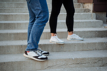 The feet of two people stand on the steps of the stairs, stylish new sneakers, modern shoes, casual style in clothes, bare ankles, white sneakers, blue jeans.