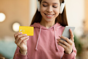 Positive teen girl in headphones using smartphone and credit card for online shopping at home, selective focus