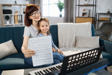 Portrait of teacher and her small student playing piano at home. Mom and daughter playing musician instrument on background of light living room at home.