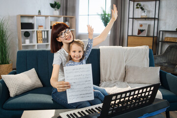 Mother teaching playing piano using notes her daughter. Little girl learning piano at home. Close...