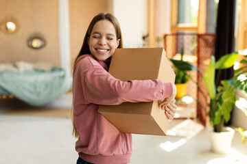 Satisfied teenager girl hugging cardboard box and smiling with closed eyes, standing in bedroom...