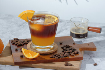cold Iced black coffee mixed with orange juice and orange slices in glass on wooden plate in white background with nature leaves silhouette shadow. two tones layer fresh summer drink in the morning
