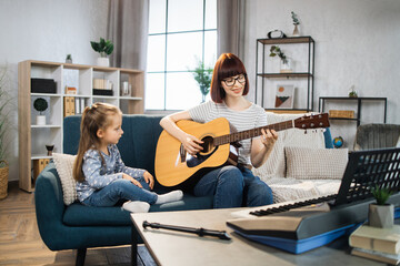 Beautiful young woman and her charming little daughter are smiling while playing acoustic guitar at...