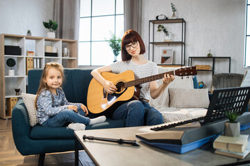Little cute girl with music teacher having lesson at guitar at school of music. Smiling young red hair woman looking at small kid girl sitting on the sofa and playing guitar together in classroom.