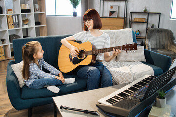Mom and little happy girl in music therapy by playing guitar on music room. Teacher helping young female pupil in guitar lesson. Relaxing at home.