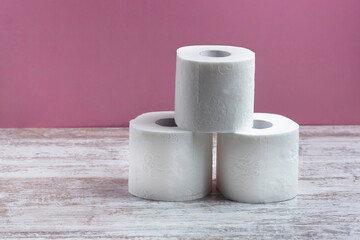 Three rolls of white toilet paper stand in a pyramid on a wooden table on a pink background