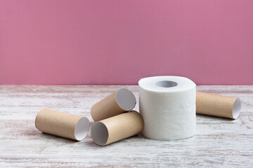 A roll of soft white toilet paper among empty toilet paper rolls.  On a wooden table on a pink background