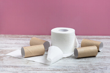An unwound roll of soft white toilet paper among empty toilet paper rolls.  On a wooden table on a pink background