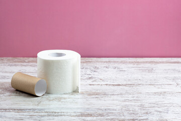 One roll of white perforated toilet paper is standing next to an empty roll.  On a wooden table on a pink background