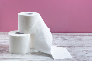 Rolls of white perforated toilet paper stand in a pile, one roll is unwound.  On a wooden table on a pink background