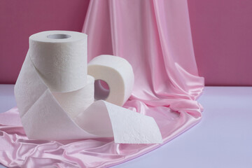 Three rolls of white perforated toilet paper, one unwound, on a pink silk background