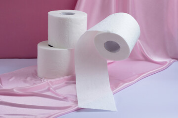 Two rolls of white perforated toilet paper stand, one flies, on a pink silk background