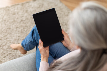 European old lady with gray hair looks at tablet with empty screen, surfing in internet in room...