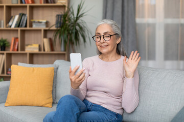 Smiling european old woman with gray hair sitting on sofa, waving hand in phone, making video call