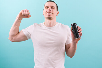 A man in a white t-shirt smiles, holds an empty black aluminum can and shows a biceps on a blue...