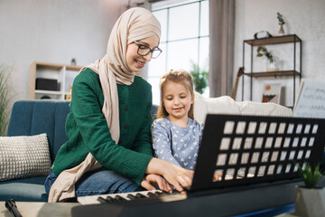 Child girl learning to play piano using notes together with her muslim mother in hijab. Remote...