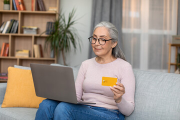 Smiling european senior woman with gray hair in glasses works on laptop and shows credit card