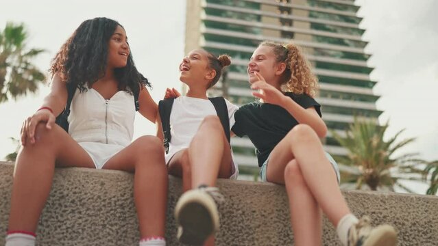 Three girls friends pre-teenage are sitting on the waterfront smiling and emotionally talking holding each other embracing. Teenagers on the outdoors in urban cityscape background
