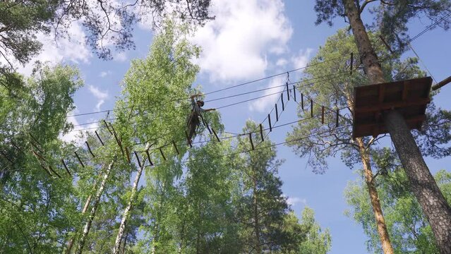 A brave teenage girl overcomes a difficult obstacle - a ladder, walking along a rope stretched between tall trees in a rope town
