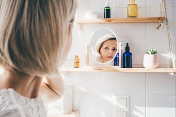 Beautiful middle-aged woman looking in the mirror in white eco friendly bathroom. Wooden shleves and reusable cosmetics bottles. Wellnes concept - obrazy, fototapety, plakaty