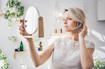 Middle-aged woman looking in the mirror in light bathroom with biophilic minimalistic design. Aging skin care. Wellnes - obrazy, fototapety, plakaty