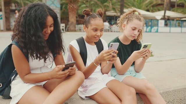 Three pre-teenage girls friends are sitting on the waterfront watching videos, photos, posting on social networks on the outdoors in urban citiscape background. Camera moves around