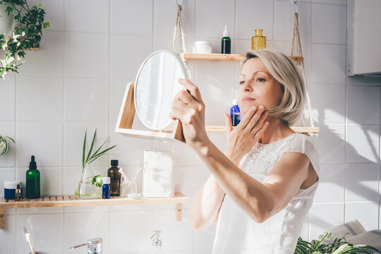 Beautiful middle-aged woman taking care of her face and looking in the mirror. White eco-friendly bathroom interior. Natural light. Wellness
