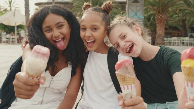 Laughing three girls friends pre-teenage are sitting on the waterfront testing ice cream. Three teenagers enjoy ice cream on hot summer day