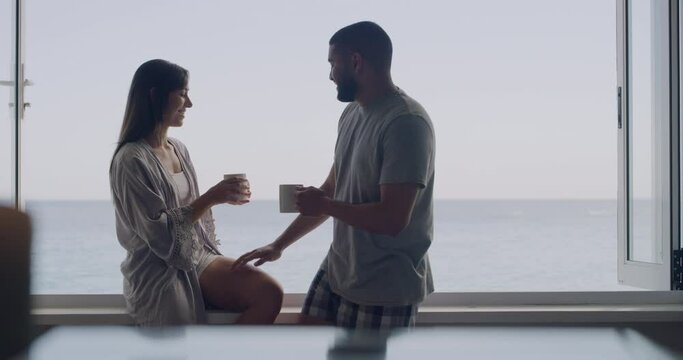 Young and diverse mixed race couple kissing and drinking coffee while sitting outside with the beach in the background. Handsome young man and his beautiful girlfriend enjoying a morning by the ocean