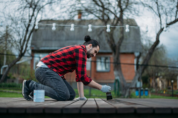 A young man in home clothes paints a wooden deck in the garden with a brush.