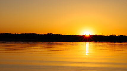 Naklejka premium Wonderful stunning orange golden sunset reflecting on water river lake. Silhouette of forest trees growing at water. Beauty of nature, landscape, horizon, skyline, summer evening, wallpaper, design.