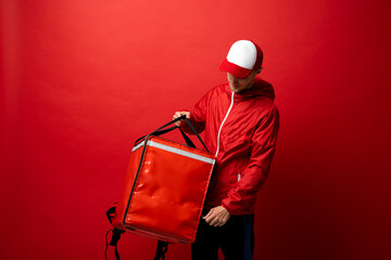 Professional delivery employee man 20s in red cap T-shirt uniform workwear work as dealer courier with red thermal food bag backpack isolated on red background. Service concept.