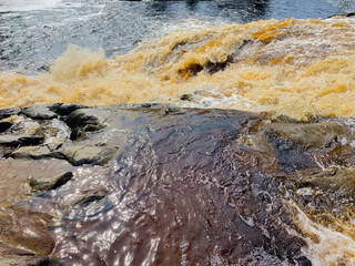 Brown-yellow water runs along large dark stones and flows into a river, which flows in a bubbling foaming stream. High quality photo