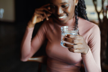 Close-up of young smiling cute African-American woman with long dark braids wearing pink roll-neck sweater, sitting at beige table, holding glass of water, looking aside. Selective focus, vertical. 