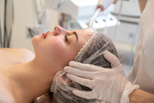 Beautician applying electric Darsonval facial massage treatment with special tool over female patients forehead in beauty salon