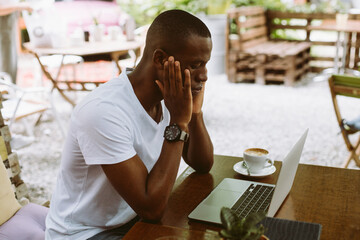 Puzzled and confused multicultural dark skin man using laptop, sitting in cafe outdoors, looking at monitor. Business and freelancing problems. Waiting results of job interview and drinking coffee.